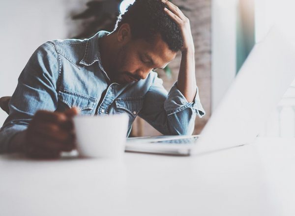 Tired man sitting at desk with his head tilted down, eyes closed. One hand holds a coffee cup, other hand props up his head