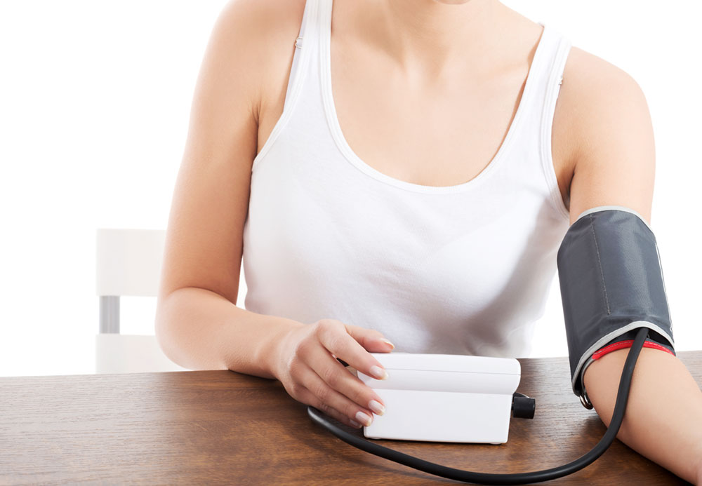 Woman in white tank top sitting at table, checking her blood pressure with the cuff around one arm and a hand on the monitor